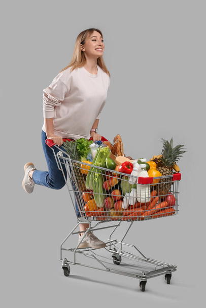 Young woman with shopping cart full of groceries on grey background - Фото, изображение