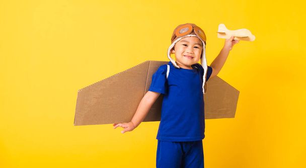 Happy Asian handsome funny child or kid little boy smile wear pilot hat play and goggles with toy cardboard airplane wings fly hold plane toy, studio shot isolated yellow background, Startup freedom - Фото, изображение