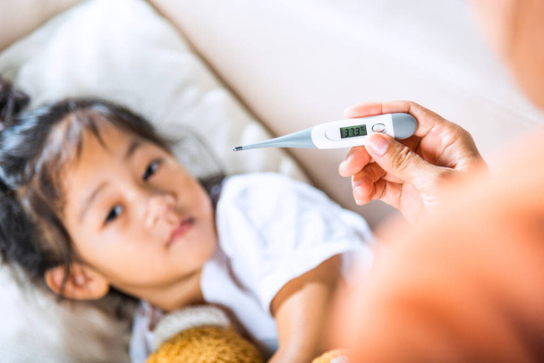 Sick kid. Mother checking temperature of her sick daughter with thermometer in mouth, child laying in bed taking measuring her temperature for fever and illness - Photo, Image