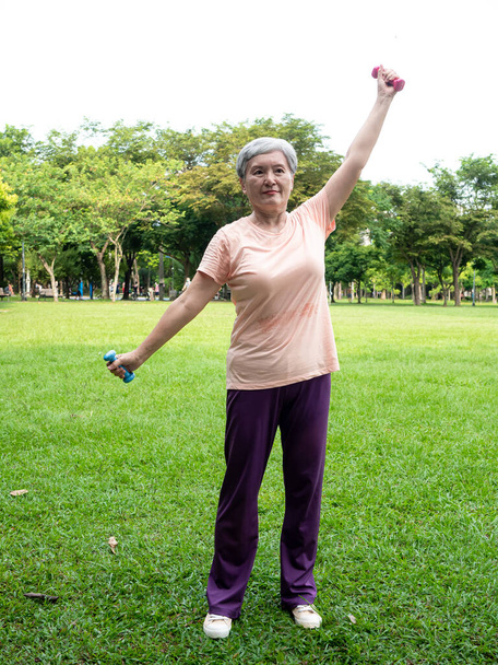 Портрет зрілої азіатки 60-х років у спортивному одязі, що працює рукою і піднімає вправи гантелі з релаксацією для здорового в парку на відкритому повітрі
. - Фото, зображення