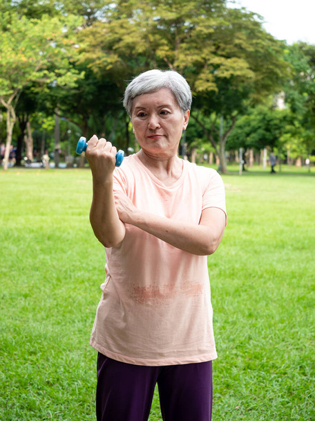 Retrato de mujer asiática madura de 60 años usando ropa deportiva haciendo ejercicio de brazo y levantando ejercicio de mancuerna con relajación para saludable en el parque al aire libre. - Foto, imagen