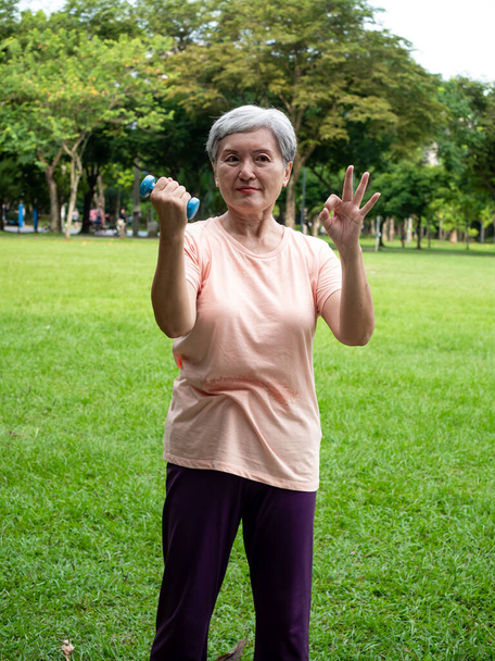 Πορτρέτο της ώριμης γυναίκας της Ασίας 60 φορώντας αθλητικά ρούχα κάνει προπόνηση βραχίονα έξω και ανύψωση αλτήρα άσκηση με χαλάρωση για υγιή στο πάρκο εξωτερική. - Φωτογραφία, εικόνα