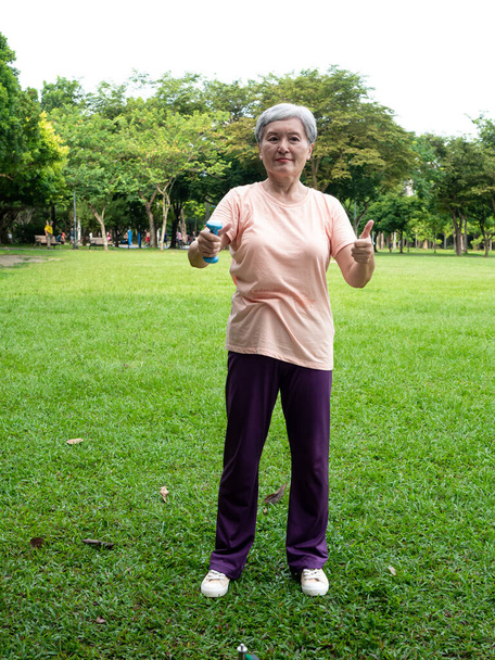 Retrato de mujer asiática madura de 60 años usando ropa deportiva haciendo ejercicio de brazo y levantando ejercicio de mancuerna con relajación para saludable en el parque al aire libre. - Foto, imagen