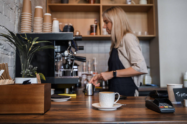 Shot of coffee cup on table and aged female barista with blond hairs. - Foto, Bild