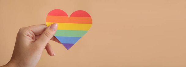 Mano femenina sosteniendo el corazón del arco iris sobre fondo beige con espacio para el texto. Concepto LGBT - Foto, imagen
