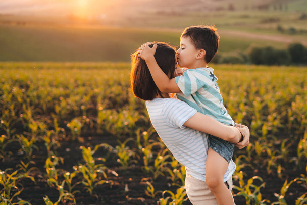 Een gelukkige jongen die zijn moeder kust op het voorhoofd, staand in hun maïsveld bij zonsondergang. Ze kijken naar hun gewassen. Gelukkige jeugd. Moeders houden ervan. Liefde tussen - Foto, afbeelding