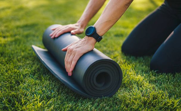 Woman hands untwist a black mat for yoga on the green grass in the summer park. Sport and healthy lifestyle concept. Relaxation practicing yoga meditation - Photo, Image