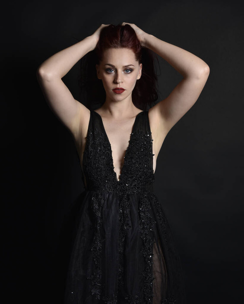 close up portrait of beautiful woman with red hair, wearing black dress with  elegant gestural hand poses on a dark studio background with moody lighting. - Fotó, kép