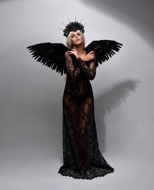 portret pięknej modelki ubranej w czarną gotycką sukienkę z nakryciem głowy i pierzastymi anielskimi skrzydłami. izolowany na tle studia. - Zdjęcie, obraz
