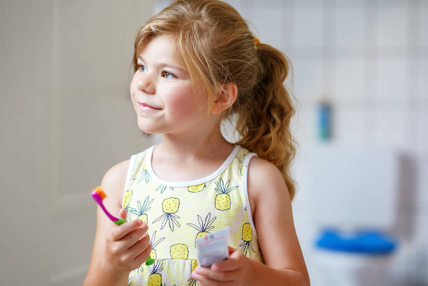 Cute little girl with a toothbrush and toothpaste in her hands cleans her teeth and smiles. Happy preschool child brushing first teeth - Zdjęcie, obraz
