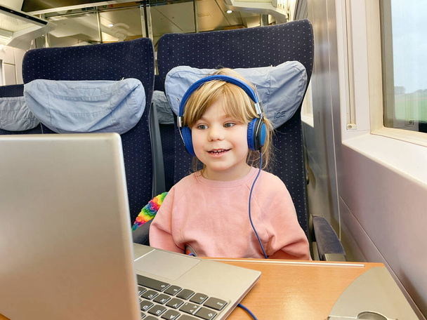 かわいい就学前の女の子は電車に座って、移動中にラップトップでテレビを見ています。鉄道で家族旅行に行く幸せな子供。ノートとヘッドフォンを持つ女の子. - 写真・画像