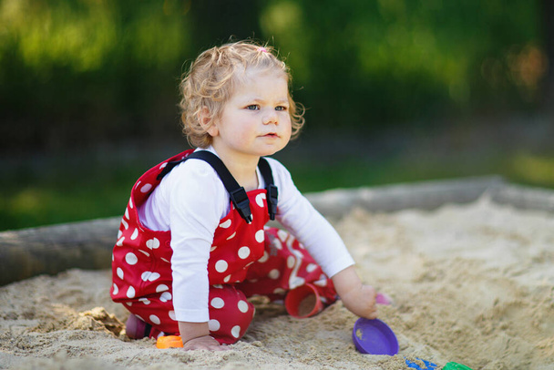 Мила дівчинка, яка грає в пісок на відкритому майданчику. Красива дитина в червоних гумових штанях розважається в сонячний теплий літній день. Дитина з різнокольоровими піщаними іграшками. Здорова активна дитина на відкритому повітрі грає в ігри
 - Фото, зображення