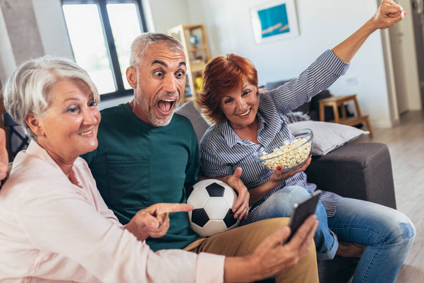 Group of senior friends support favorite sports team together use smartphone app to online bet - Photo, Image