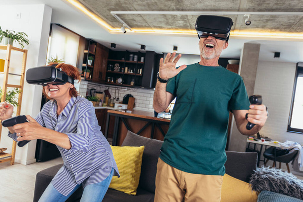 Ryhmä keski-ikäisiä ystäviä hymyilee onnellinen pelaa videopeli VR lasit kotona. - Valokuva, kuva
