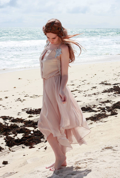 портрет красивої молодої жінки з довгим волоссям у простій сукні, що тече у вітрі, елегантна поза. океанський пляжний фон з золотим освітленням заходу сонця
. - Фото, зображення