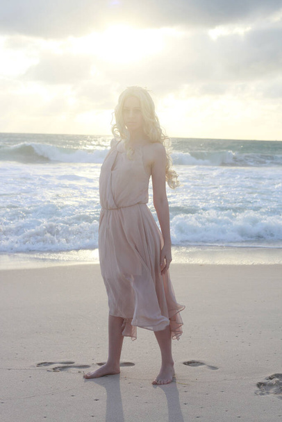 полноразмерный портрет красивой молодой женщины с длинными волосами в простом платье, текущем ветром, элегантная поза. океанский пляж фон с золотым освещением заката. - Фото, изображение