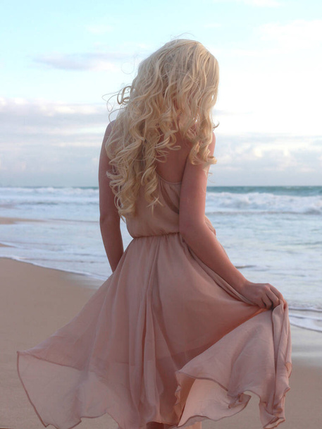 風に流れるシンプルなドレスを着て長い髪を持つ美しい若い女性の完全な長さの肖像画、エレガントなポーズ。黄金の夕焼けの海の背景. - 写真・画像