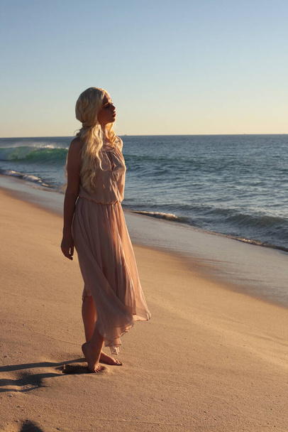Uzun saçlı, rüzgarda süzülen basit bir elbise giyen güzel bir genç kadının tam boy portresi. Zarif bir duruş. Okyanus sahili arka planında altın günbatımı ışığı. - Fotoğraf, Görsel