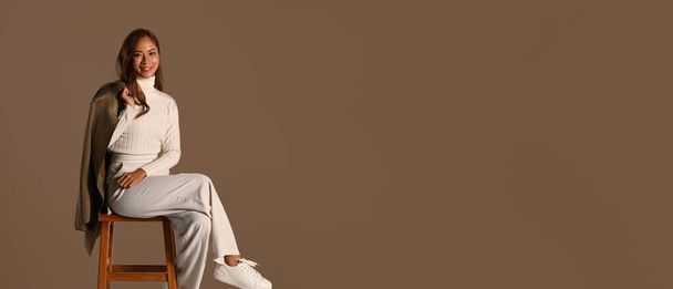 Panoramic shot of stylish woman holding trench coat sitting on wooden chair isolated on brown background. Autumn fashion concept. - Fotoğraf, Görsel