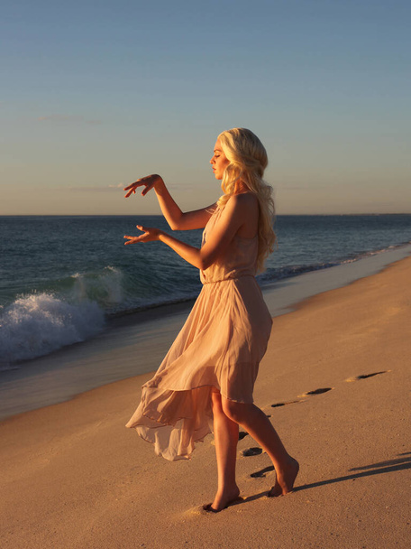 полноразмерный портрет красивой молодой женщины с длинными волосами в простом платье, текущем ветром, элегантная поза. океанский пляж фон с золотым освещением заката. - Фото, изображение