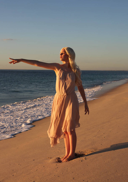 pełnowymiarowy portret pięknej młodej kobiety z długimi włosami noszącej prostą sukienkę płynącą na wietrze, elegancką pozę. ocean plaża tło ze złotym słońcem oświetlenie. - Zdjęcie, obraz