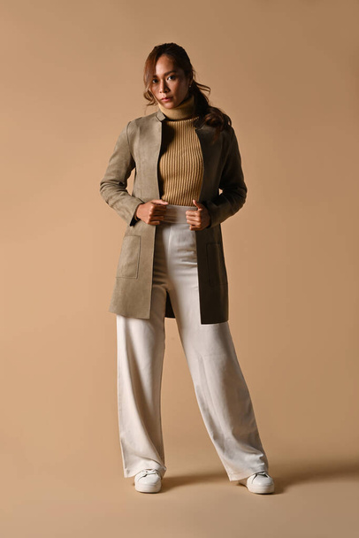 Full length portrait of fashionable woman in trench coat standing on beige background. Autumn fashion and beauty concept. - 写真・画像