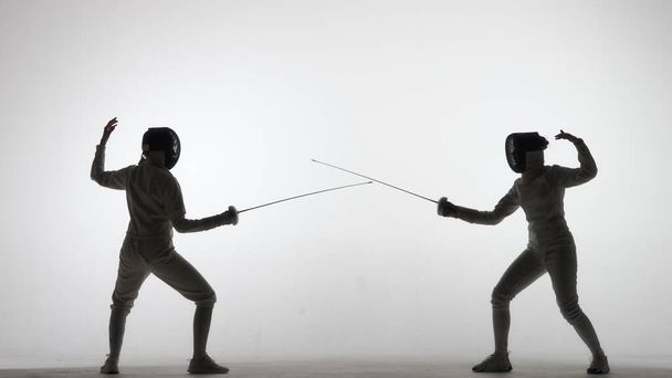 Side view of two young female swordswoman with crossed rapiers. Athletes in uniforms and protective helmet mask fighting a duel in dark studio on white background. Girls holding epees in their hands - Fotoğraf, Görsel