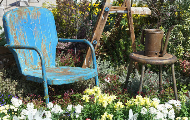 Ruosteinen vanha sininen tuoli keskellä vehreyttä ja kukkia aurinkoisessa puutarhassa                                - Valokuva, kuva