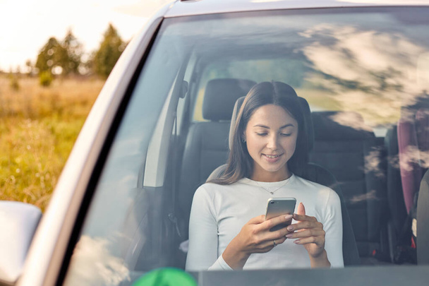 Smiling beautiful young adult woman looking at her mobile phone while sitting inside car, attractive dark haired female wearing white shirt in her automobile surfing internet via smart phone. - Foto, Imagen