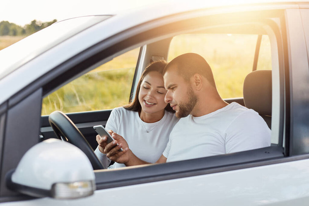 Portrait of man and woman in a new car, traveling together, wife holding smart phone and showing something to her husband, family spending nice time together while traveling. - Foto, imagen