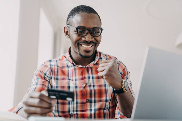 African american guy holding credit card makes yes gesture using e banking service at laptop. Black man satisfied by successful financial transaction with cashback. Online banking, e commerce. - Foto, immagini