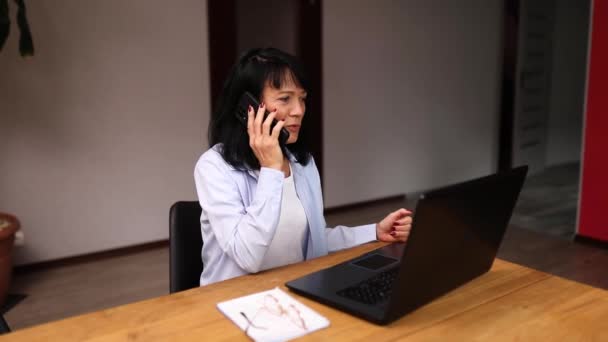 年上の女性従業員は、クライアントまたはビジネスパートナーとスマートフォンコール携帯電話を持っています。シニア女性はノートパソコンで職場で電話で話しています. - 映像、動画