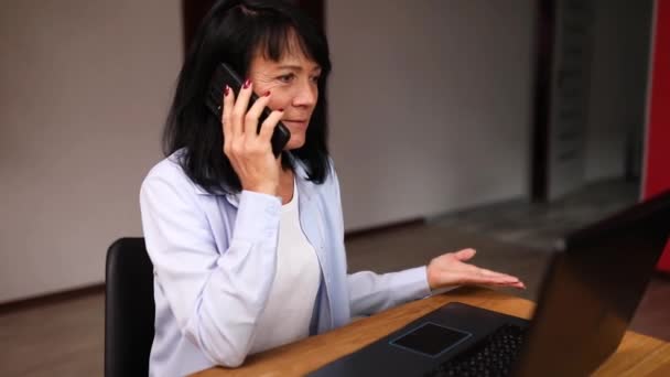 年配の女性が職場でノートパソコンで電話で話している。年上の女性従業員は、クライアントやビジネスパートナーとスマートフォンコール携帯電話を持っています. - 映像、動画