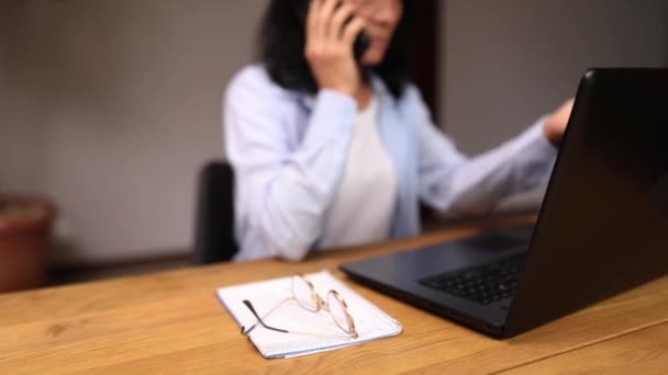 Starší zaměstnankyně mají chytrý telefon s klientem nebo obchodním partnerem. Senior žena mluví po telefonu na svém pracovišti s notebookem. - Záběry, video