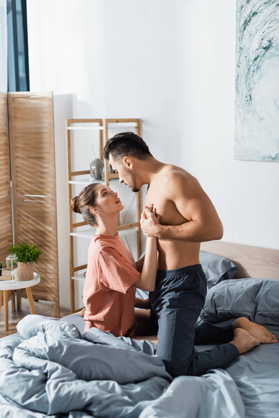 passionate woman touching muscular torso of boyfriend kneeling on bed - Photo, Image