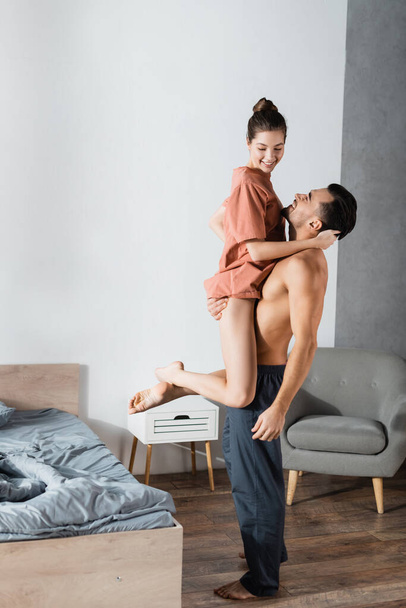 hombre musculoso en pantalones de pijama sosteniendo novia sonriente en el dormitorio - Foto, imagen