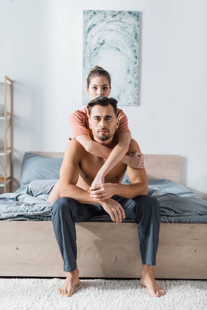 young woman looking at camera while embracing shirtless man sitting on bed in pajama pants - Photo, Image