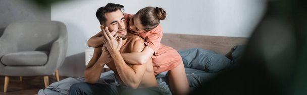 passionate woman embracing shirtless man lying on bed on blurred foreground, banner - Foto, imagen