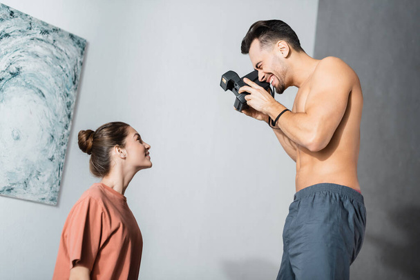 zijaanzicht van vrolijke shirtloze man met vintage camera fotograferen van vriendin in t-shirt - Foto, afbeelding