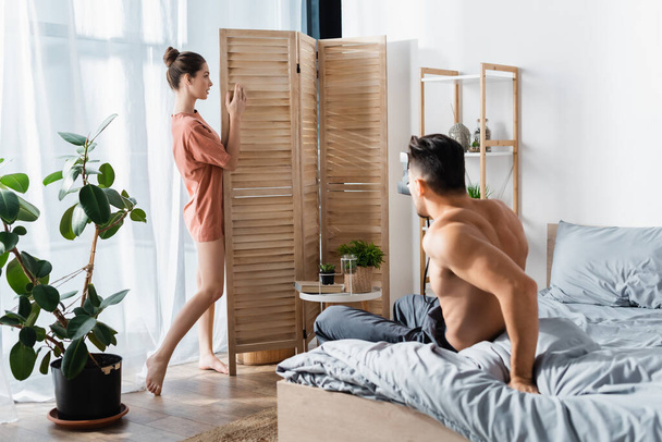 sexy woman in t-shirt posing near room divider and shirtless man with vintage camera in bedroom - Photo, Image