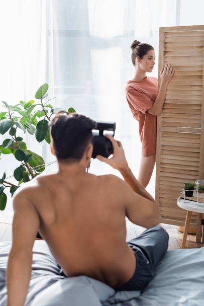 donna seducente posa vicino divisorio stanza mentre l'uomo senza maglietta con fotocamera vintage scattare foto su primo piano sfocato - Foto, immagini