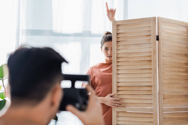 mujer joven apuntando hacia arriba con el dedo mientras posando cerca del divisor de habitación y novio borroso con cámara vintage - Foto, Imagen