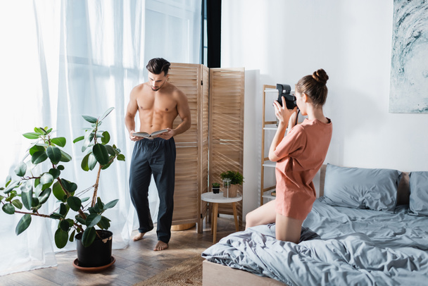 muscular man in pajama pants posing with book near girlfriend with vintage camera - Photo, Image