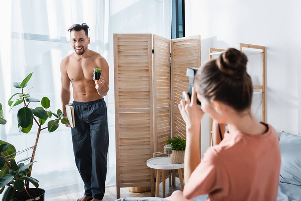 cheerful shirtless man with book and flowerpot posing near blurred girlfriend with vintage camera - Fotoğraf, Görsel