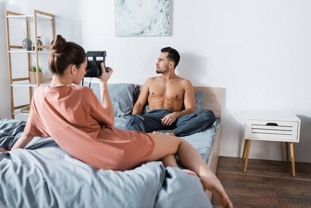 sexy man with muscular torso sitting on bed near young girlfriend with vintage camera - 写真・画像
