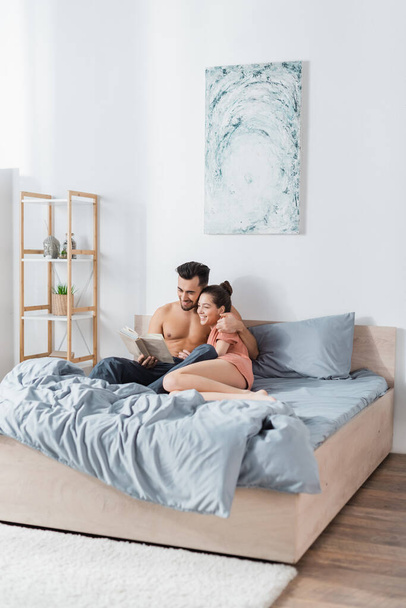 cheerful couple sitting on grey bedding and reading book - Foto, immagini