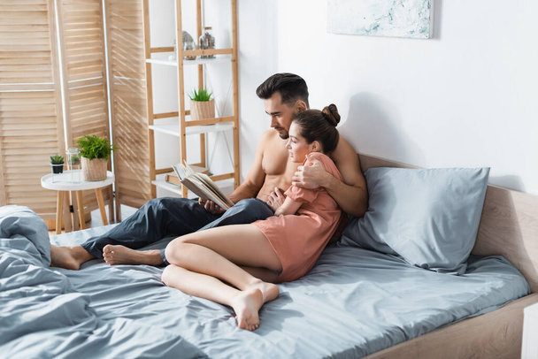 young shirtless man embracing girlfriend in t-shirt while reading book in bedroom - Valokuva, kuva