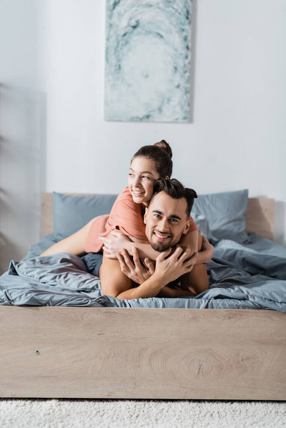 happy man looking at camera near girlfriend hugging him in bedroom - Zdjęcie, obraz