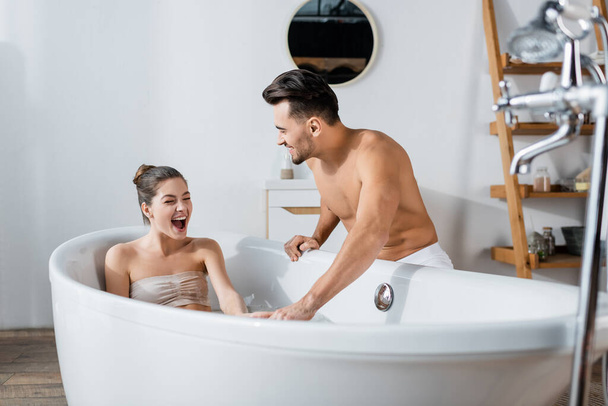 взволнованная женщина с открытым ртом, принимающая ванну рядом с парнем без рубашки - Фото, изображение