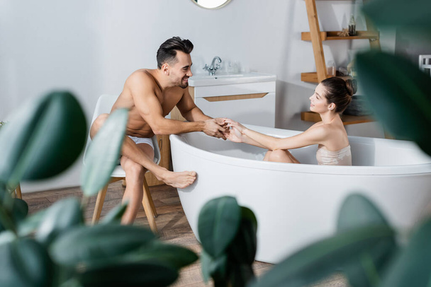 sensual young woman holding hands with sexy and smiling boyfriend while taking bath on blurred foreground - Фото, изображение